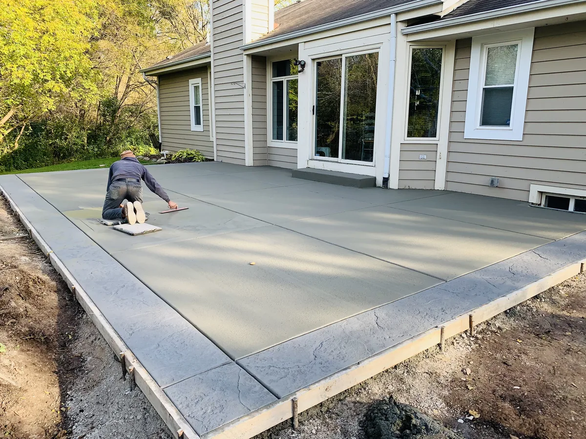 Worker troweling the surface of a freshly poured concrete patio at the back of a house